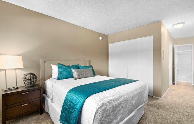 Mar Vista CA Apartments - Vista - Furnished Bedroom with Plush Carpeted Floors, Bed, Wooden Nightstand with Drawers and a Lamp, Beige Walls, and Large White Closet