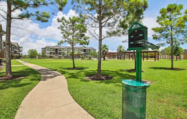 A green trash can is in the foreground of a grassy area with a pathway and trees.