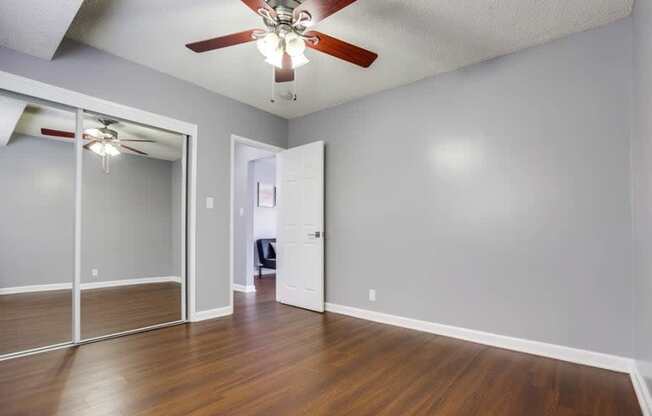 Tuscany Villas Apartments in West Covina - Bedroom with Hardwood Flooring, Mirrored Sliding Door Closet, Window, Ceiling Fan, Popcorn Ceiling, Grey Walls, and Access to Hallway