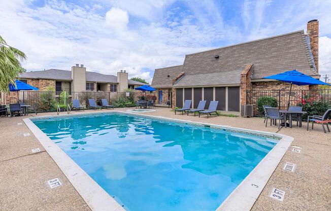 A clear swimming pool surrounded by lounge chairs and tables, with blue umbrellas, set against a backdrop of brick buildings under a partly cloudy sky.
