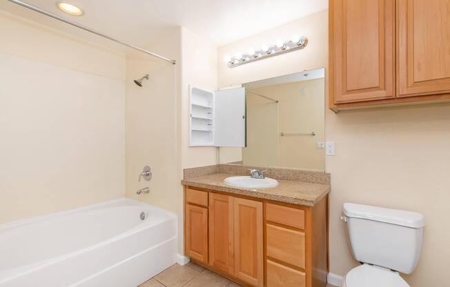 A clean, well-lit bathroom featuring a white bathtub, a wooden vanity with a sink, a mirror above the sink, and a medicine cabinet. The walls are painted a soft color, and there is a toilet nearby. The overall design is modern and simple, with tile flooring.