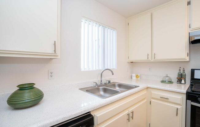 Bright kitchen featuring a two-basin stainless steel sink, light-colored cabinets, and a countertop made of a white speckled material. A green decorative vase sits on the counter, and a window with vertical blinds allows natural light to illuminate the space. Modern appliances and organized decor complete the inviting atmosphere.