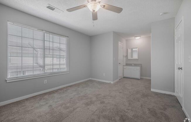 A bright, empty room with light gray walls and beige carpet. It features a ceiling fan, a large window with blinds, and a corner that includes a bathroom vanity area with a mirror. Natural light filters into the space, creating an inviting atmosphere.