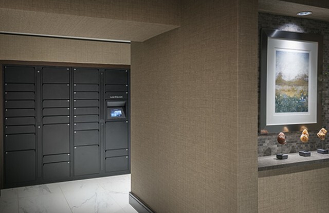An around-the-corner glimpse of the package room, with marble floors, gray lockers of various sizes installed along the wall, and a touch-screen access panel.