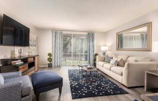 A living room with a grey chair, a blue ottoman, a white couch, a wooden coffee table, a flat screen TV, and a large mirror.