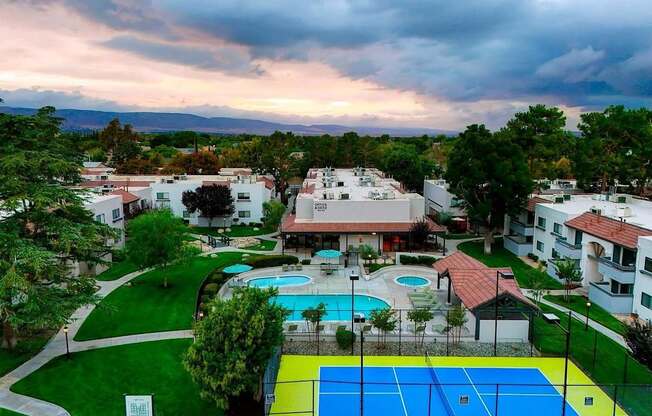 A tennis court is surrounded by a pool and apartment buildings.