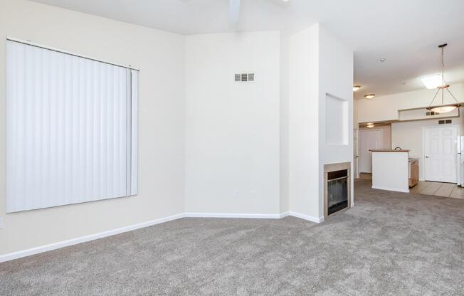 A spacious living area featuring beige walls and light gray carpet. A large window with vertical blinds on the left, a small fireplace on the right, and a view of an adjacent kitchen area in the background. The space is well-lit and open, creating a welcoming atmosphere.