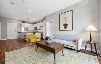 A living room with hardwood-style flooring, natural light, high ceilings, and open access to the kitchen with a breakfast bar.