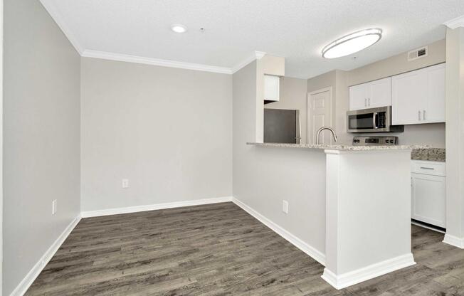 A modern kitchen and dining area with gray walls, dark vinyl flooring, and a bar counter. The kitchen features white cabinets, a stainless steel microwave, and a black refrigerator. Soft overhead lighting illuminates the space, creating a bright and inviting atmosphere.