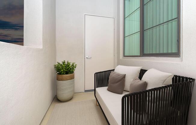 A cozy balcony space featuring a modern black woven sofa with two cushions, a floor rug, and a large potted plant. A closed door and a large window with green-tinted glass are visible, enhancing the serene atmosphere. The walls are painted white, creating a minimalist aesthetic.