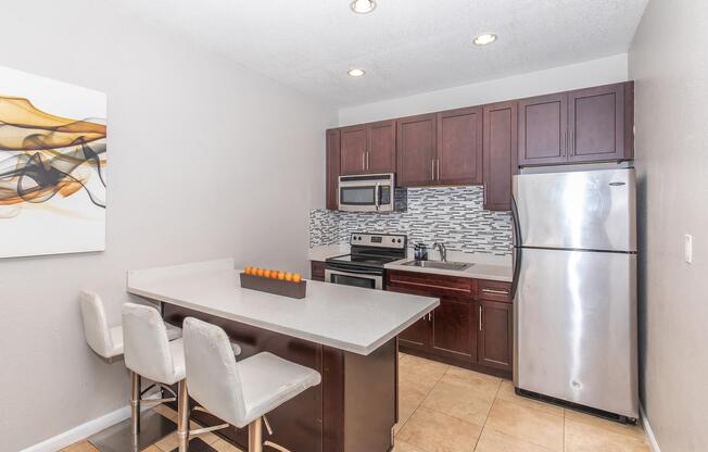 Modern kitchen featuring dark wood cabinets, stainless steel appliances including a microwave and refrigerator, gray quartz countertops, and a minimalist dining area with barstools. Wall art is displayed, adding a contemporary touch. Overall, the space is bright and inviting with neutral tones.