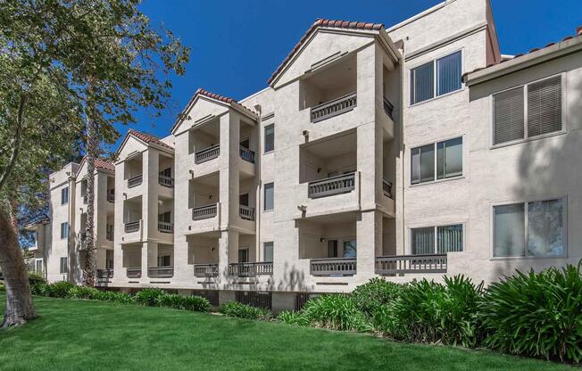 A two-story apartment building with a stucco exterior and multiple balconies. The building is set against a bright blue sky and features green grass and shrubs in the foreground. The architecture includes arched windows and a tiled roof, conveying a modern yet classic style.