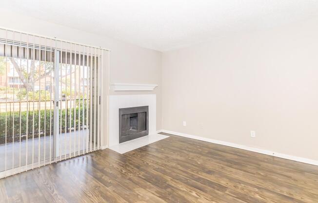 A light and airy living room featuring a large window with vertical blinds, a white fireplace, and hardwood-style flooring. The walls are painted in a soft beige color, creating a neutral and inviting atmosphere. The space is unfurnished, emphasizing its open layout.