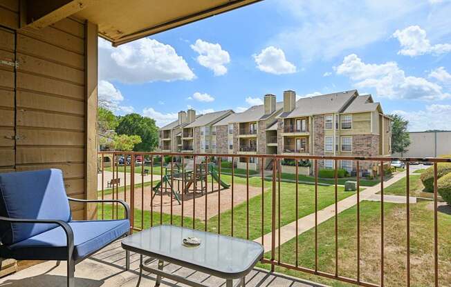 A balcony with a blue chair and table overlooks a grassy area and buildings.