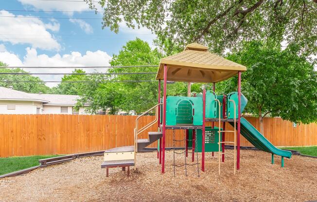 Colorful children's playground equipped with slides, climbing structures, and play panels. It sits on a bed of wood chips, surrounded by a wooden fence and green trees, under a partly cloudy sky. The playground invites outdoor play and exploration.