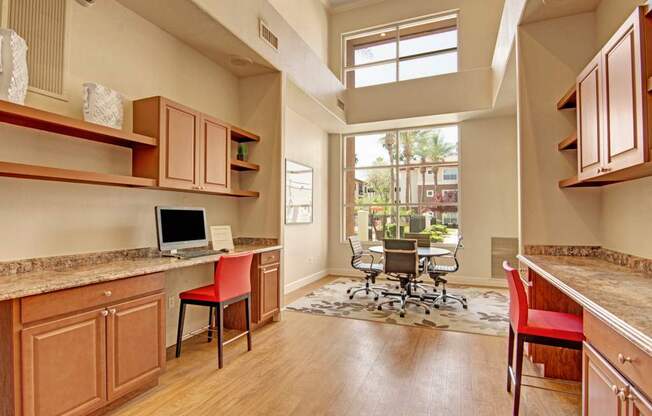 A modern business center here at Mozaic at Steele Park featuring built-in wood cabinetry, granite-style countertops, and dedicated computer workstations with comfortable seating. Tall windows and elevated ceilings fill the space with natural light, while a collaborative table area overlooks the beautifully landscaped courtyard, creating a productive and inviting environment for work or study.