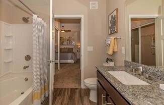 A spacious bathroom featuring a bathtub with a shower curtain, a modern sink with a granite countertop, and a towel rack. In the background, a bedroom is visible with a large bed and decorative artwork on the walls. The room has neutral colors and contemporary design elements.