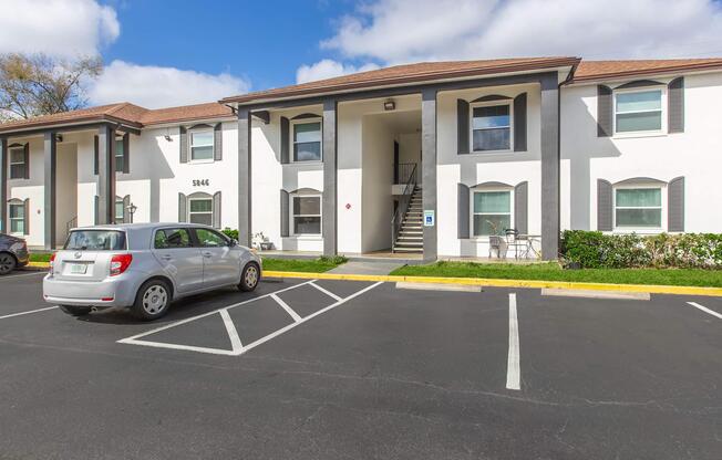 Exterior view of a two-story residential building with a white facade and brown roof. A silver car is parked in the foreground, with several parking spaces visible. The entrance features a staircase, and there are green shrubs and a small seating area outside the building. Bright blue sky with some clouds in the background.