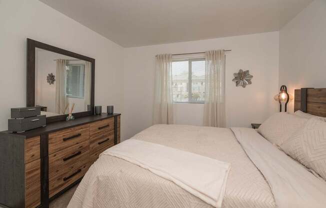 A bedroom with a large bed, a wooden dresser, and a window with curtains at Sierra Glen Apartments, California, 95621
