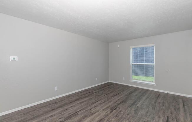 Empty room with light gray walls and wood laminate flooring. A single window with blinds is visible, allowing natural light to enter. There is a thermostat on the wall, but no furniture or decorations in the space, creating a minimalist and spacious atmosphere.