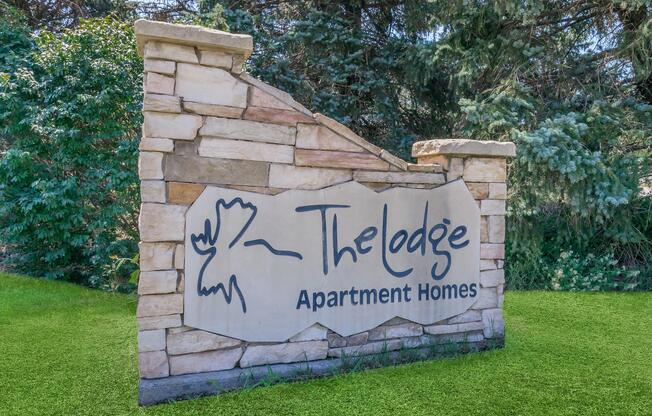 Sign at The Lodge Apartment Homes featuring a stone structure with a stylized logo and text. Surrounded by greenery, the sign presents a welcoming entrance to the apartment complex.