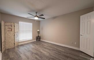 Spacious empty room with light gray walls, a wooden floor, and a ceiling fan. A decorative room divider is visible on the left, and a small lamp on a table is positioned near the window. Natural light streams through the window, which has white blinds. The door is closed in the background.