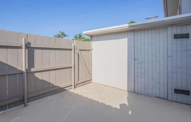 A small, enclosed patio or outdoor area features a light-colored concrete surface. The space is bordered by a wooden fence, with one side having a gate. In the background, a white wall and the clear blue sky can be seen, along with some palm trees visible over the fence.