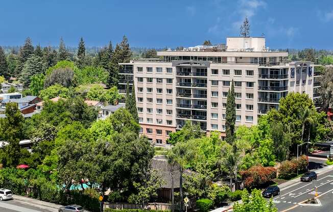 A large building with a flat roof is surrounded by trees and is located on a street with cars.