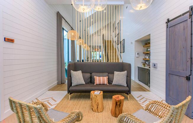 Modern living room featuring a gray sofa with a decorative pillow, two woven chairs, and wooden side tables. The space is bright with large windows, neutral-toned walls, and pendant lighting. A wooden staircase and open kitchen area are visible in the background, creating a cozy, inviting atmosphere.