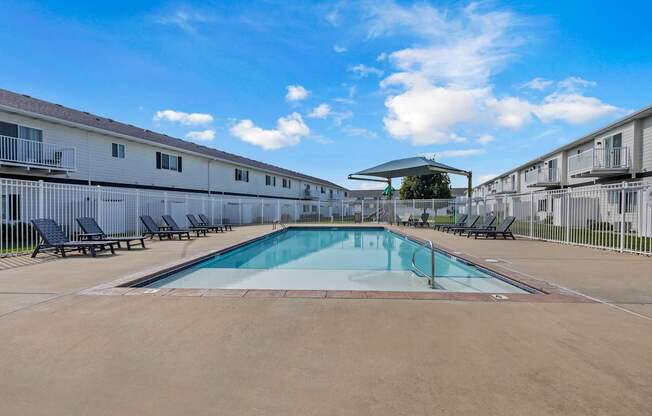 A swimming pool in a sunny day surrounded by chairs and buildings.
