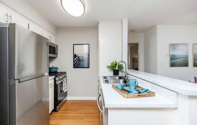 A modern kitchen featuring stainless steel appliances, including a refrigerator and stove. The countertop is made of white speckled material, with a tray holding a blue cup and small plant. A framed picture hangs on the wall, and the space is well-lit with natural light from an adjacent room.