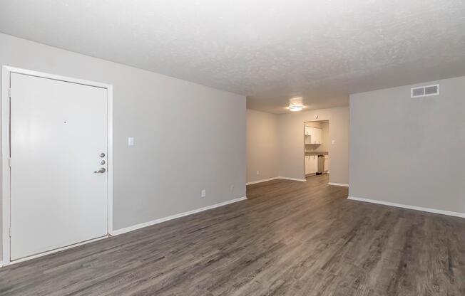 Empty living room space with gray walls and a light-colored floor. A white door is visible on the left, leading outside. In the background, there's a glimpse of a kitchen area. The room has a clean, modern look with ample natural light.