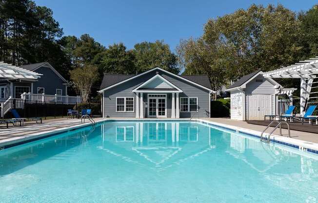 A swimming pool in front of a house with a deck and lounge chairs.