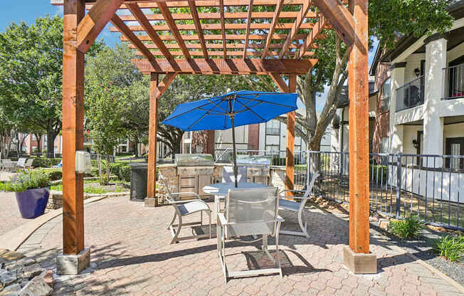 A wooden pergola with a blue umbrella is in the foreground of a brick patio.