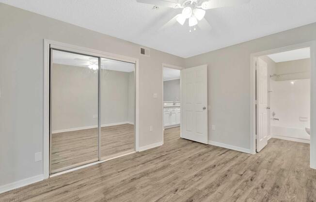 A spacious room with light gray walls, ample natural light, and a ceiling fan. There is a mirrored closet on one wall, and an open door leading to a bathroom area visible in the background. The flooring is a warm wood laminate, creating a cozy atmosphere.