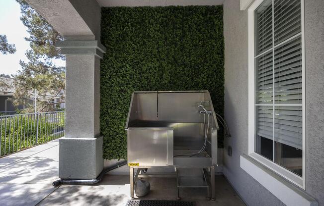 A stainless steel outdoor sink placed in a shaded corner of a patio, surrounded by green artificial foliage. The sink features a faucet and a drain, with a concrete surface beneath it. Sunlight filters through nearby trees, illuminating the area.