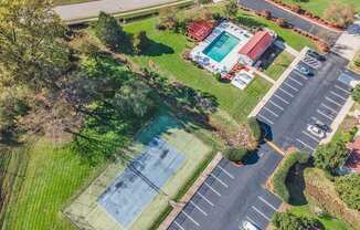 A tennis court is surrounded by a parking lot and a grassy area.