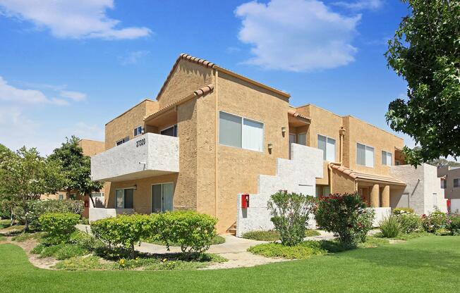 A two-story apartment building with a light beige exterior and white accents. The structure features a tiled roof, large windows, and landscaped greenery around it, including shrubs and small trees. The sky is bright blue with a few clouds, creating a cheerful atmosphere.