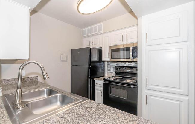 Modern kitchen featuring a granite countertop, stainless steel sink, black refrigerator, and black oven/stove. The cabinets are white with a classic design, and the kitchen is well-lit with a round overhead light.