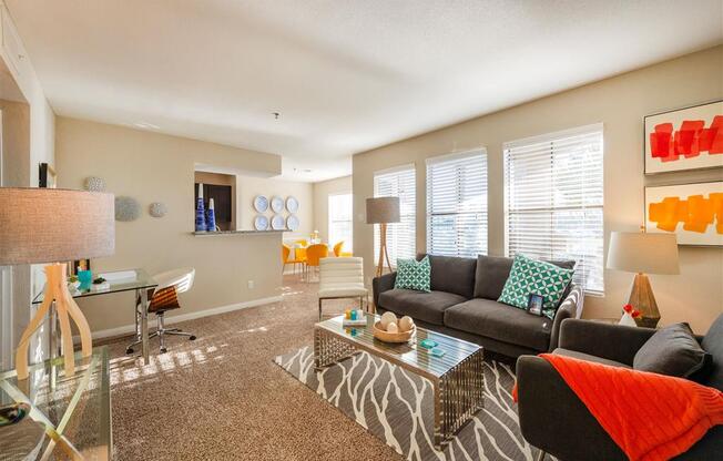 League City, TX Apartments - Anchor at South Shore - Furnished Living Room with Carpet Flooring, Large Windows with Blinds, Stylish Decor, and a Dining Area Near the Kitchen