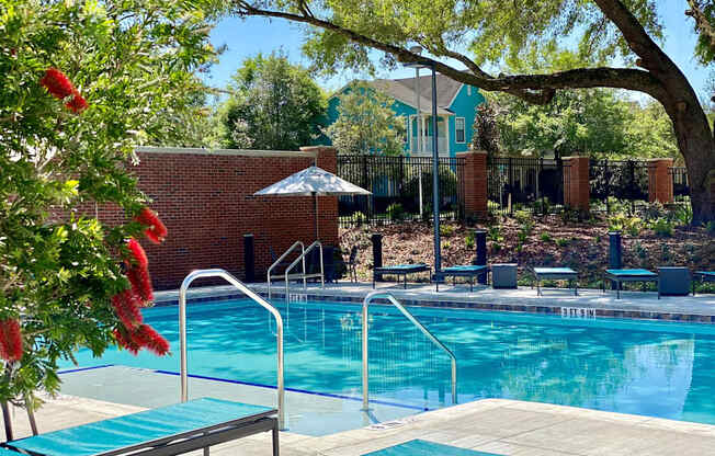 A beautiful gated swimming pool surrounded by lounge chairs, an umbrella, a large tree, and well-maintained landscaping.