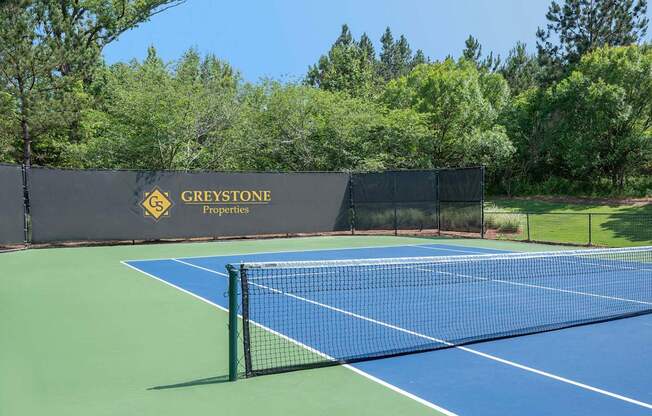 A tennis court with a blue surface and a black fence with the words "Greystone Properties" written on it.