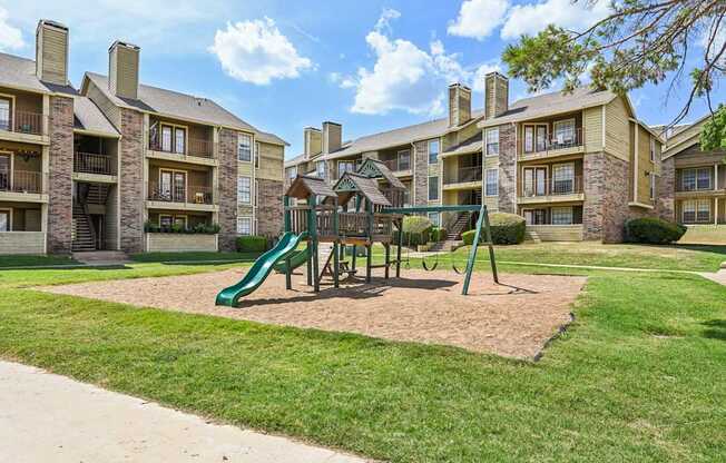 A playground with a green slide and a brown sandbox in front of apartment buildings.