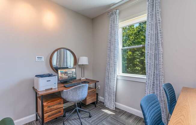 The Residences at Crosspoint study nook in the cyber lounge shows a wooden desk with a printer and desktop computer, a round wall mirror above, a gray upholstered swivel chair, and a floor lamp beside a curtained window looking out onto trees.