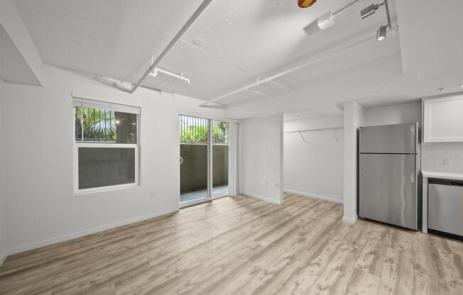 A spacious, empty room with light wood flooring, white walls, and large windows. It features a sliding glass door leading to an outdoor area, a modern stainless steel refrigerator, and overhead lighting. The layout is open, creating a bright and inviting atmosphere.
