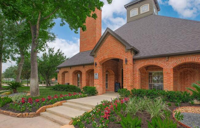 A charming brick building with a pitched roof and a tall chimney, surrounded by lush green landscaping. Colorful flowerbeds line the walkway leading to the entrance, framed by trees and shrubs. The sky is partly cloudy, adding to the inviting atmosphere of the property.