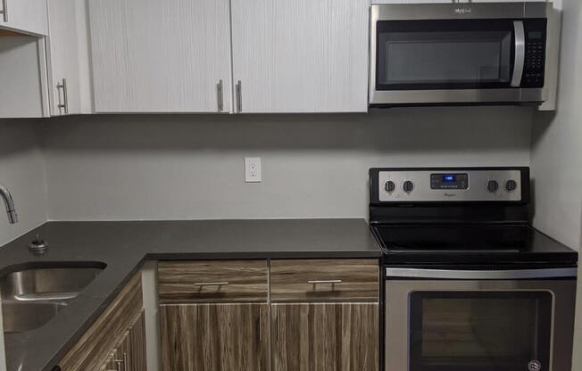 Image of kitchen, there are dark wood-like cabinets below with white cabinets on top. There are black countertops and stainless steel appliances. There is a sink to the left and an oven with a microwave above on the right. The walls are a light grey.