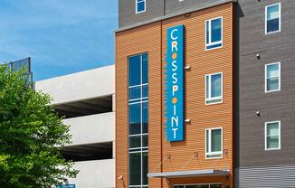 The Residences at Crosspoint apartment community at 42 Wellman Street in Lowell, MA features an orange corrugated-metal façade with a tall blue vertical sign reading "Crosspoint" mounted on the building next to a white concrete parking garage.