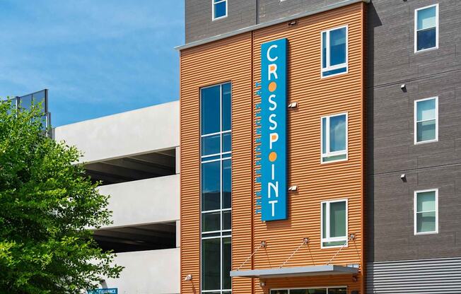 The Residences at Crosspoint apartment community at 42 Wellman Street in Lowell, MA features an orange corrugated-metal façade with a tall blue vertical sign reading "Crosspoint" mounted on the building next to a white concrete parking garage.