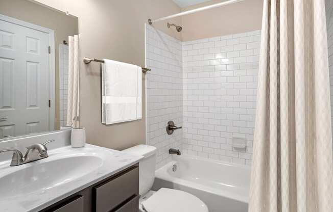 A vacant renovated bathroom with a single sink with marble countertops, gray ash cabinets below, brushed nickel plumbing fixtures, a single mounted mirror, one toilet, a towel bar, and a tub/shower combo with white subway tile surround.